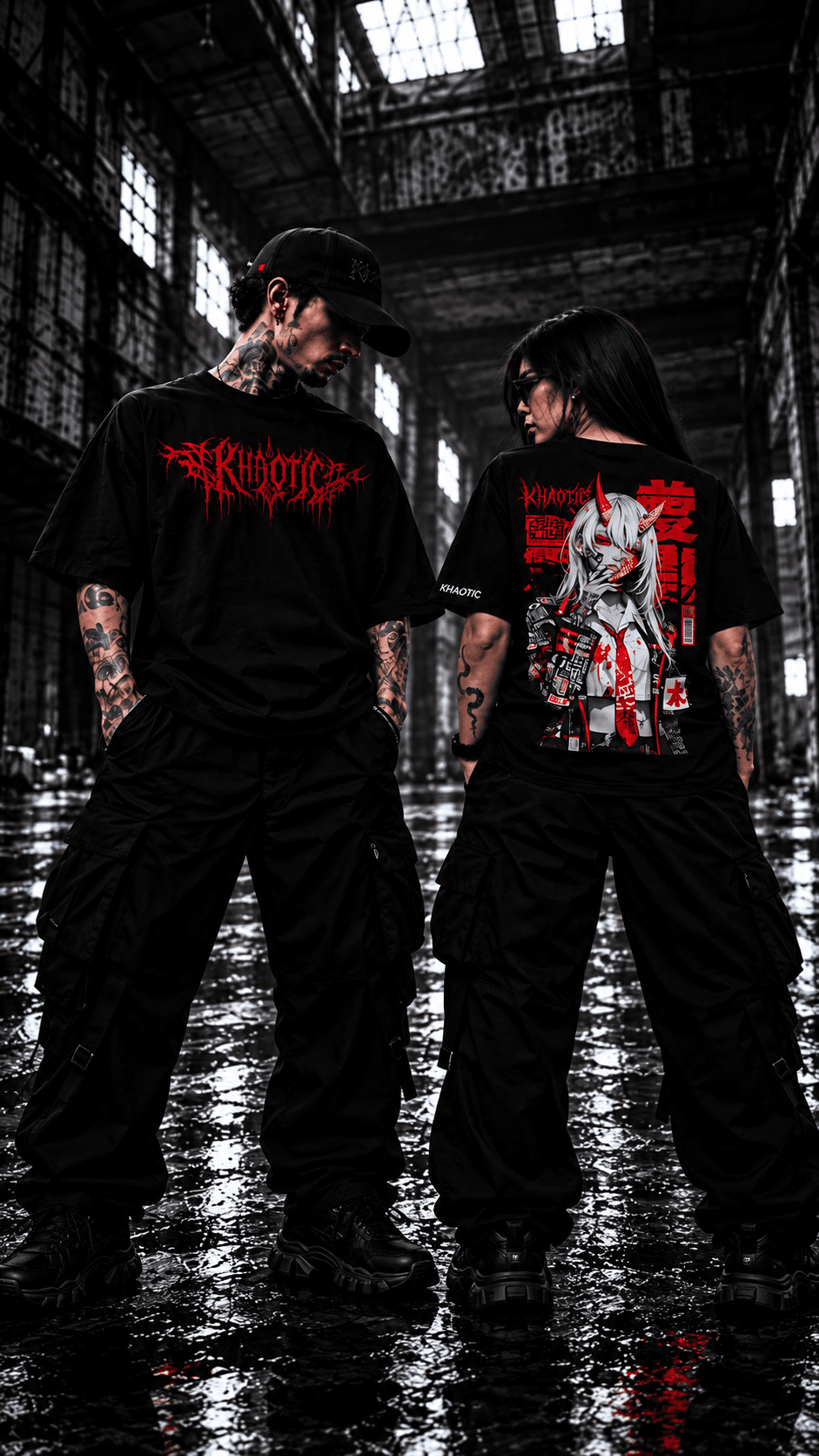 KHAOTIC man facing forward and woman seen from behind, both wearing black graphic T-shirts in an industrial warehouse